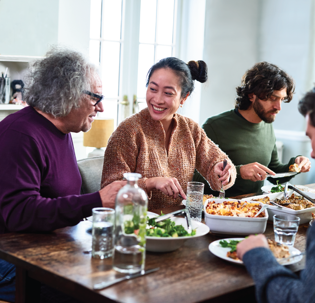 Group of mixed aged relatives sitting at dining table enjoying home cooked fall meal, talking and laughing.