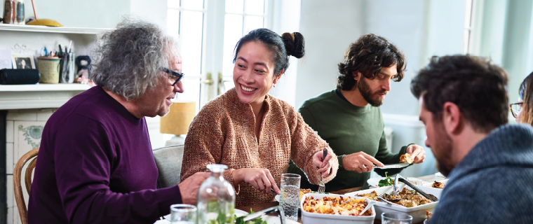 Group of mixed aged relatives sitting at dining table enjoying home cooked fall meal, talking and laughing.