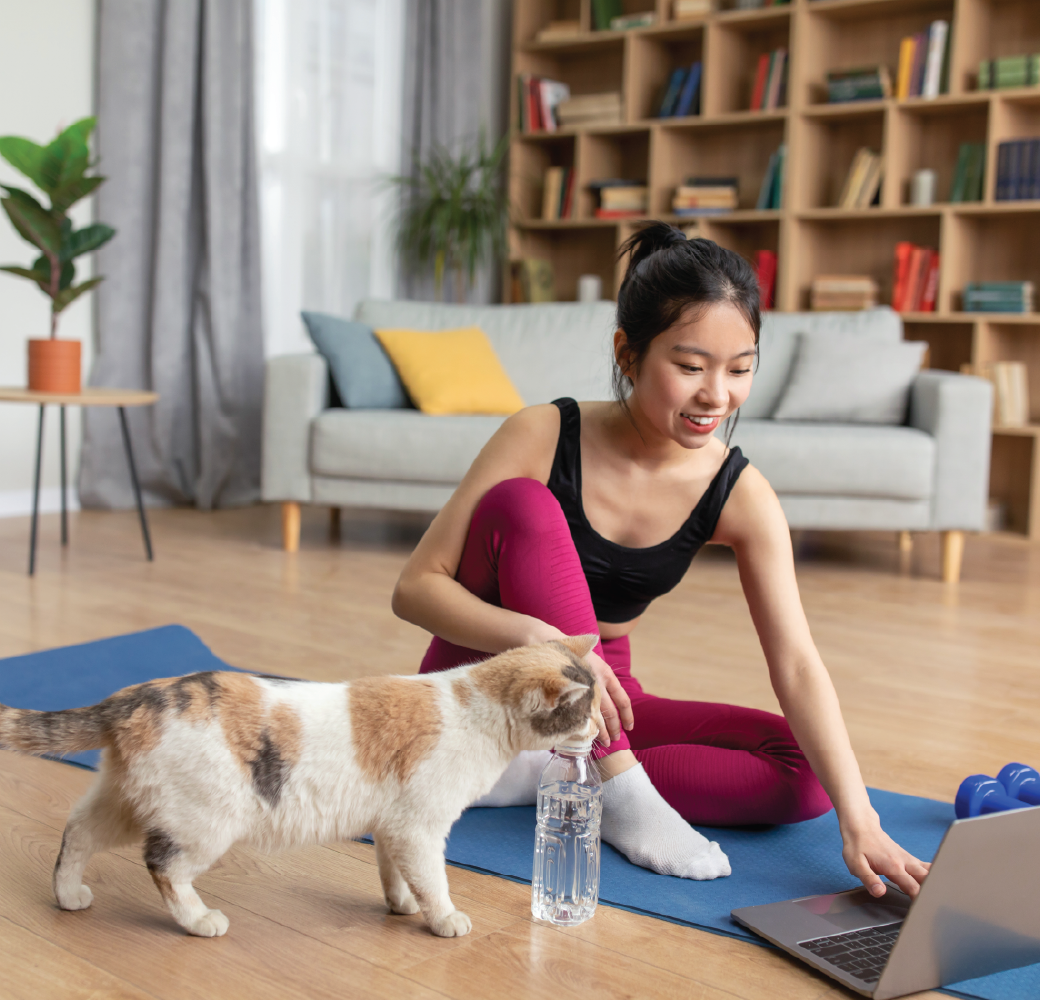 Cat and female owner exercising together at home. Sporty asian lady watching online training tutorial while sitting on mat near her pet, having morning yoga.