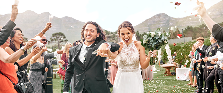 Man wearing suit and woman wearing wedding dress walking up grassy wedding aisle while having petals thrown at them by guests